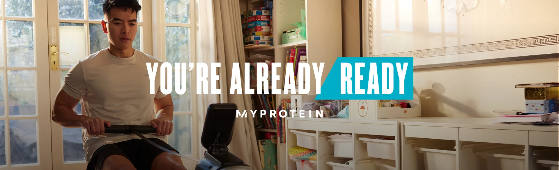A man workout out on a rowing machine at home with the words 'You're Already Ready - MYPROTEIN' overlaying.